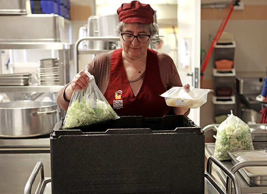 Operatrice di mensa con grembiule rosso prepara pasti confezionando verdure fresche e alimenti in una cucina professionale